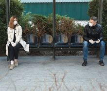 Frau und Mann sitzen mit Abstand und Manske und Abstand an einer Bushaltestelle